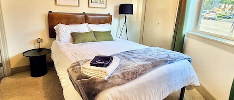 A hotel room with a bed, a nightstand, a lamp, a window with a view, and two framed pictures on the wall.
