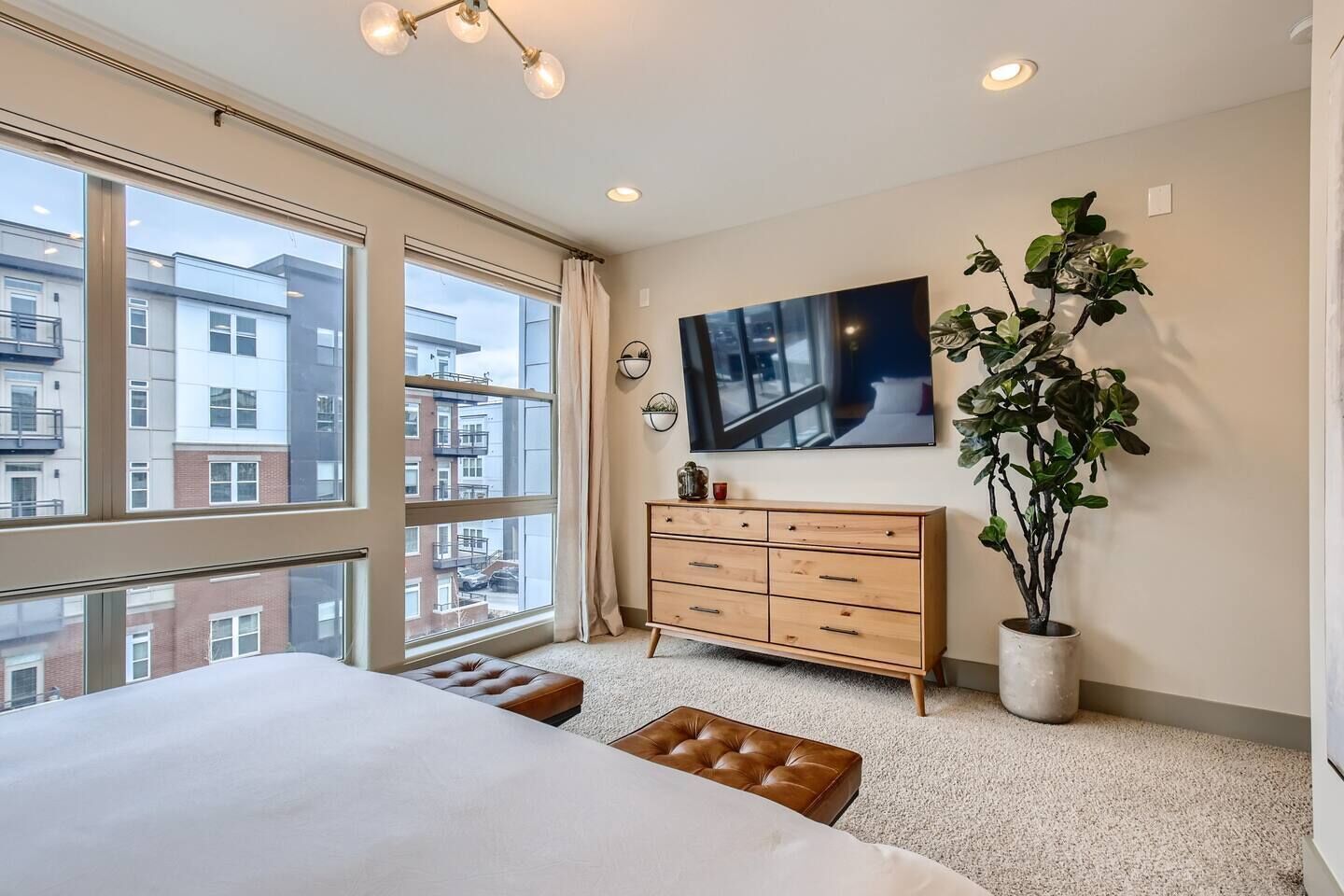 A hotel room with a large bed, a wooden dresser, a potted plant, a flat-screen TV, and a view of the city through large windows.