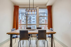 Een eetruimte met een tafel en stoelen, grote ramen voorzien van gordijnen, en uitzicht op een woongebouw.