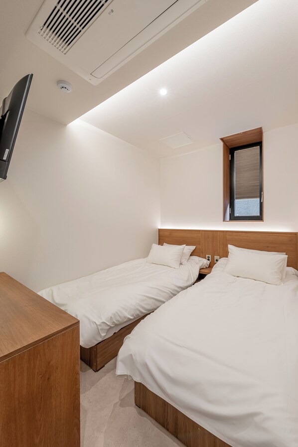Une chambre d’hôtel avec deux lits, un téléviseur, une bouche d’aération au plafond et une petite fenêtre.