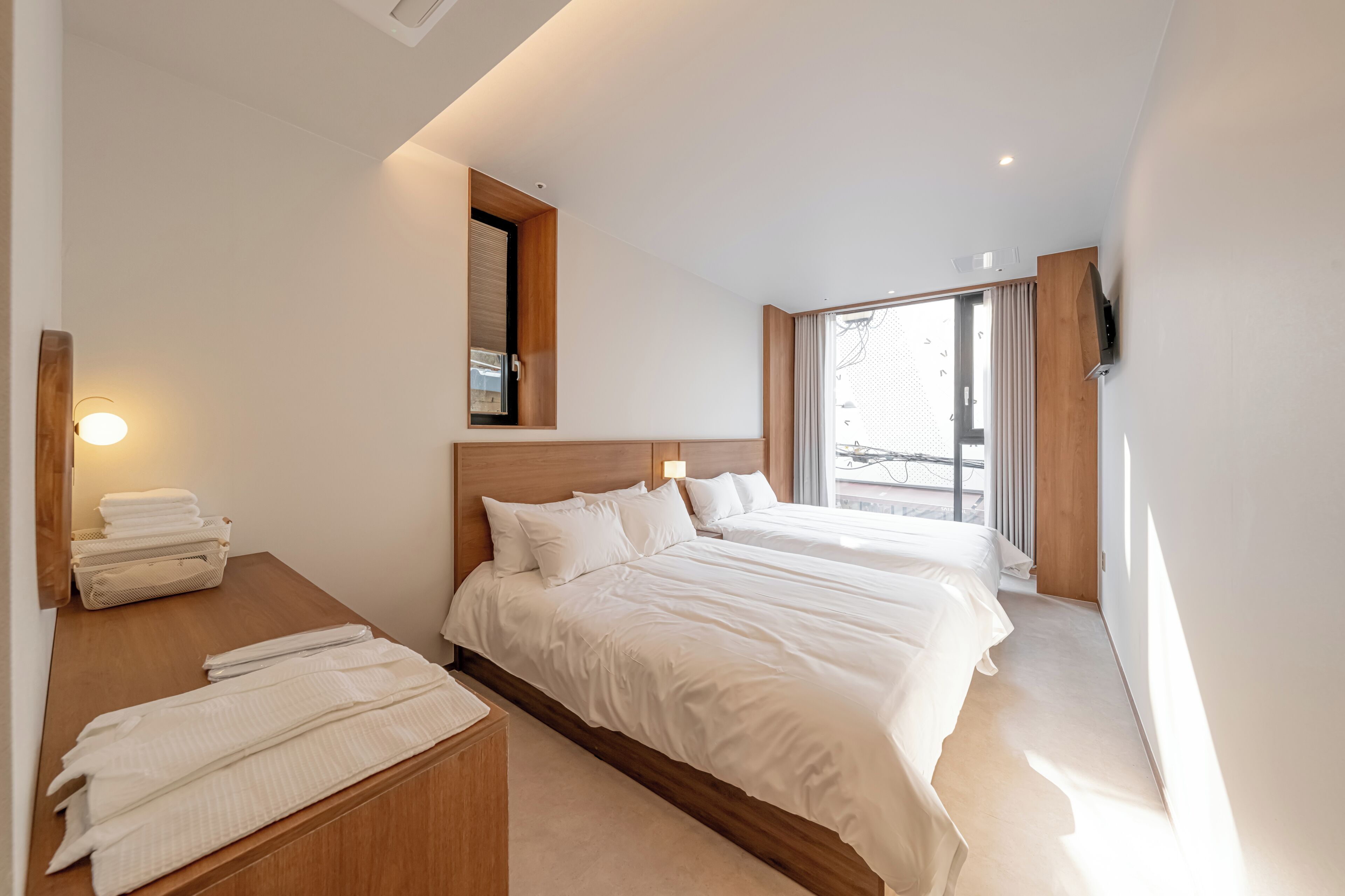 Ein modernes Hotelzimmer mit einem großen Bett, einem Holzkopfteil, einem Nachttisch mit Handtüchern und einem großen Fenster mit Vorhängen.