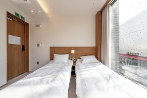 Une chambre d’hôtel avec deux lits simples, une tête de lit en bois, une petite table de chevet, un réveil fixé au mur, une fenêtre donnant sur un immeuble et une porte avec un panneau indiquant la sortie de secours en cas d’incendie.