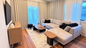 Ein modernes Wohnzimmer mit einer Couch, einem Couchtisch, einem Fernseher und einem Fenster mit Vorhängen.