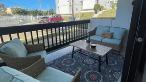 Un balcone con mobili in vimini, un tappeto decorato e vista su una strada con edifici e un'auto rossa.