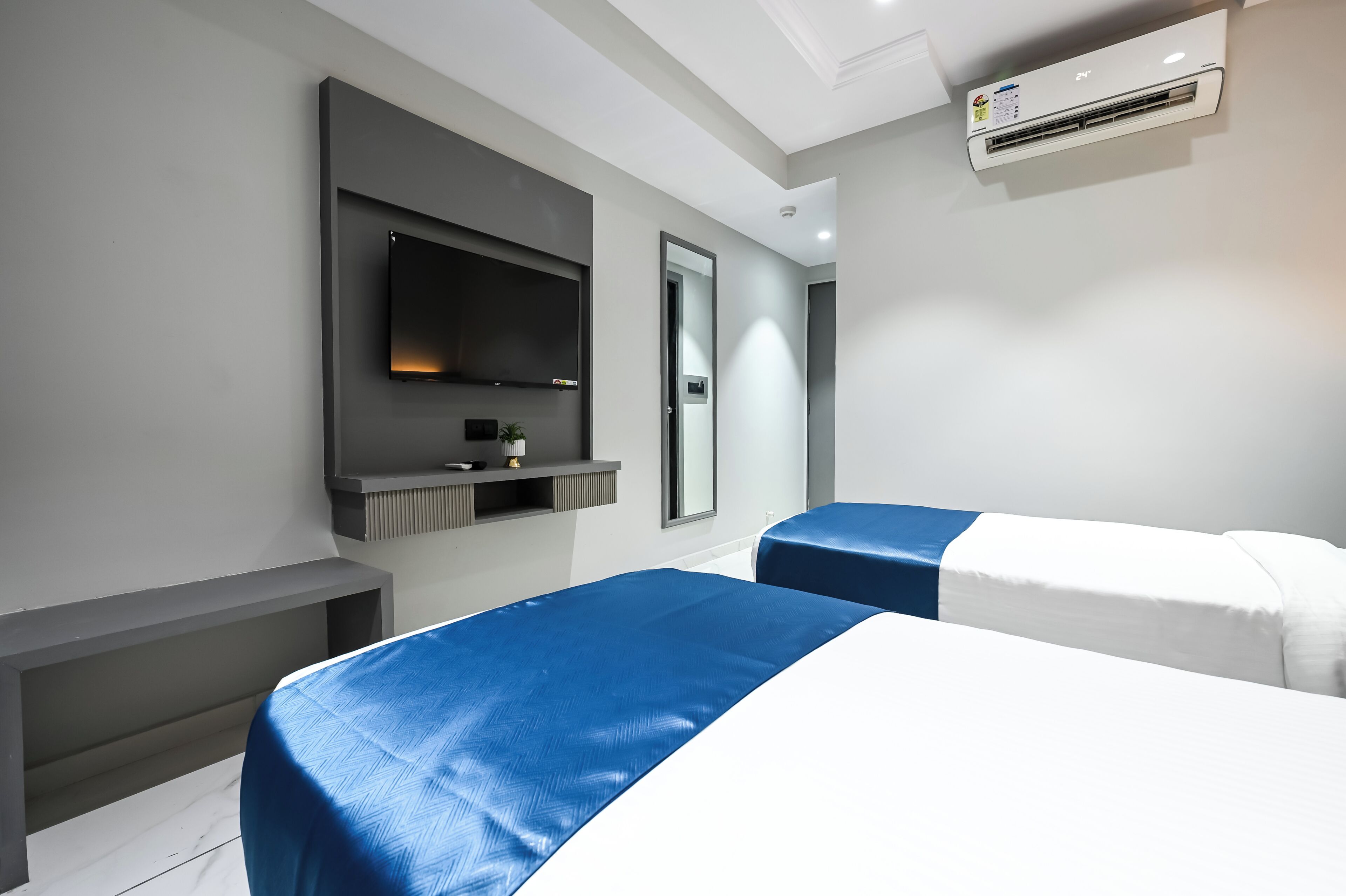 A modern hotel room with a flat-screen TV mounted on the wall, a built-in bench, and two beds with blue and white bedding.