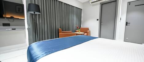 A hotel room with a bed, a chair, a table, an air conditioning unit, and a door.