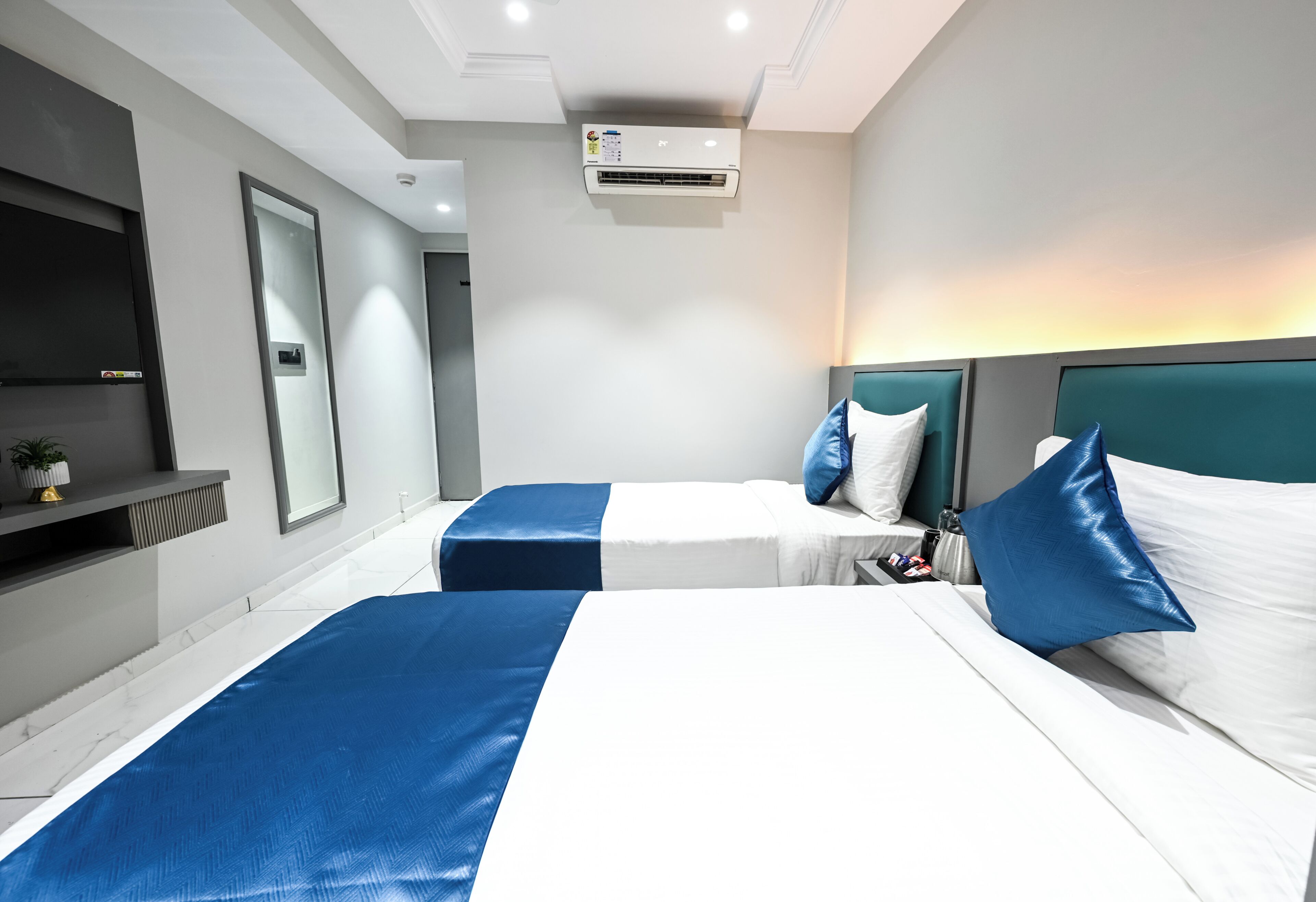A hotel room with two beds, a TV, a wall-mounted air conditioner, and a small plant.