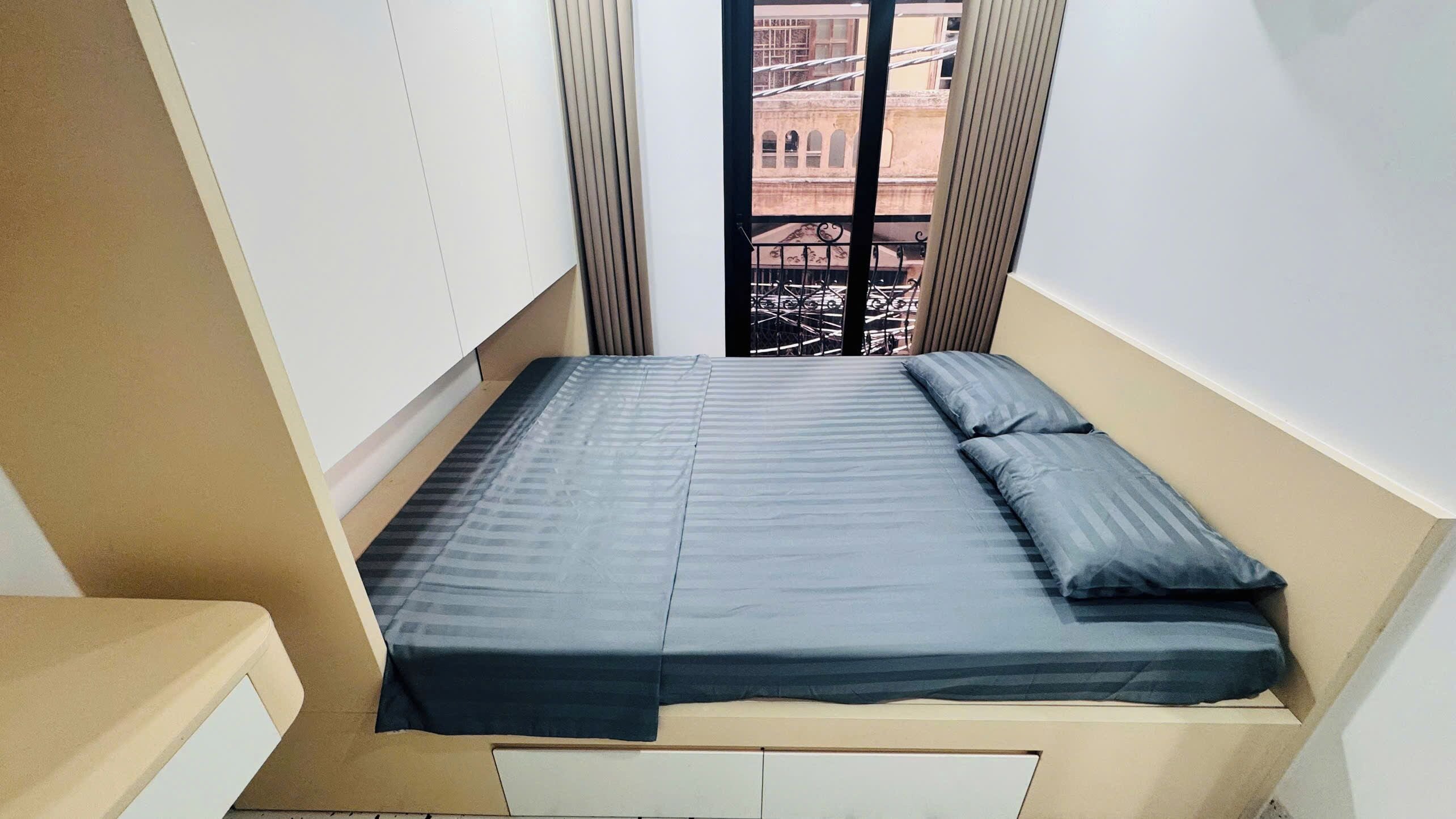 A small, narrow room with a single bed, a window with a view of a building, and a small table.