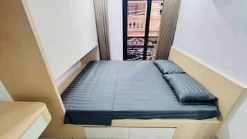 A small, narrow room with a single bed, a window with a view of a building, and a small table.