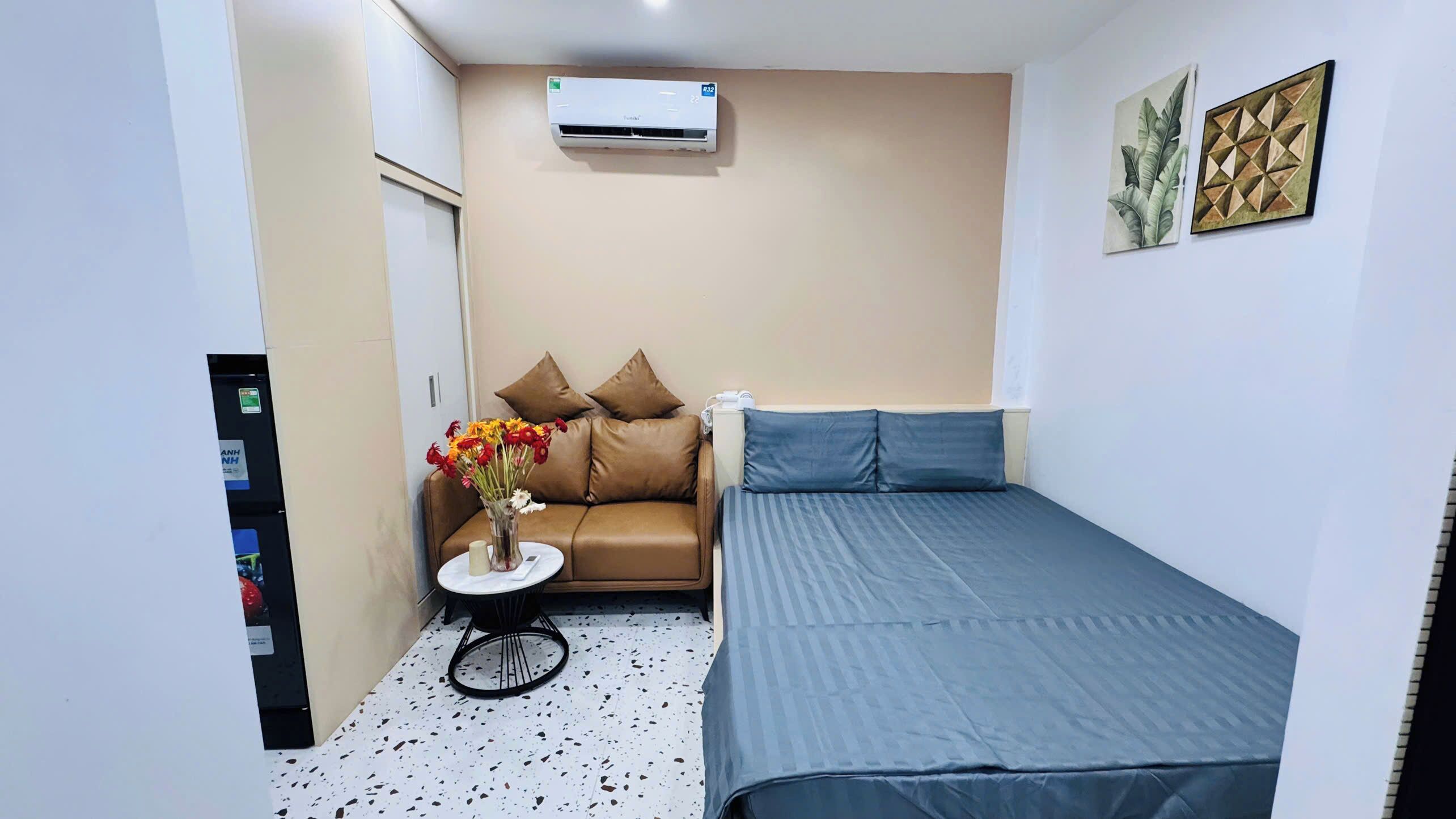 A small, compact room with a bed, a sofa, a small table with a vase of flowers, an air conditioning unit, and a wall-mounted artwork.