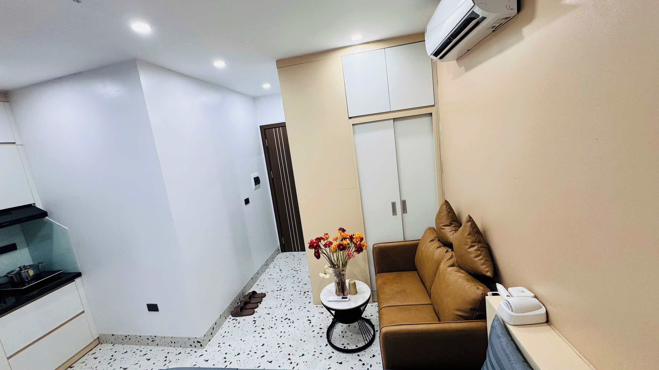 A compact living space with a kitchenette, a sofa with cushions, a small round table with a vase of flowers, and a white wardrobe.
