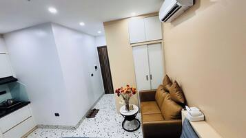 A compact living space with a kitchenette, a sofa with cushions, a small round table with a vase of flowers, and a white wardrobe.