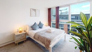 Un dormitorio moderno con una cama grande, mesitas de noche, un radiador y vistas a la ciudad a través de amplios ventanales.