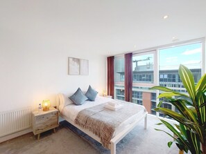 A modern bedroom with a large bed, bedside tables, a radiator, and a view of the city through large windows. - Two Bedroom Duplex Pent House in ExCeL (Greater London)