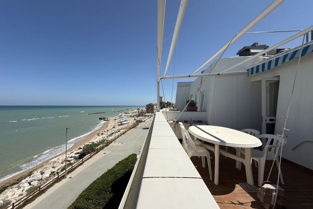 A balcony with a view of the beach, a table and chairs, and a building with a blue-striped awning.