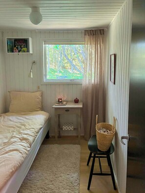 A bedroom with a bed, a desk with a lamp, a window with a view of trees, and a door.