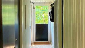 Un pasillo estrecho con pisos de madera, paredes blancas y vista a un árbol desde una puerta.