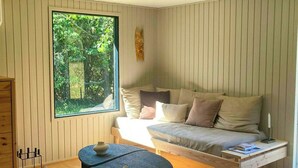Una habitación con piso de madera, un sofá, una silla, una mesita y una ventana con vista a la vegetación.