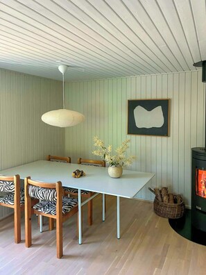 A dining area with a white table, zebra-patterned chairs, a hanging light fixture, a framed abstract artwork, and a wood-burning stove.