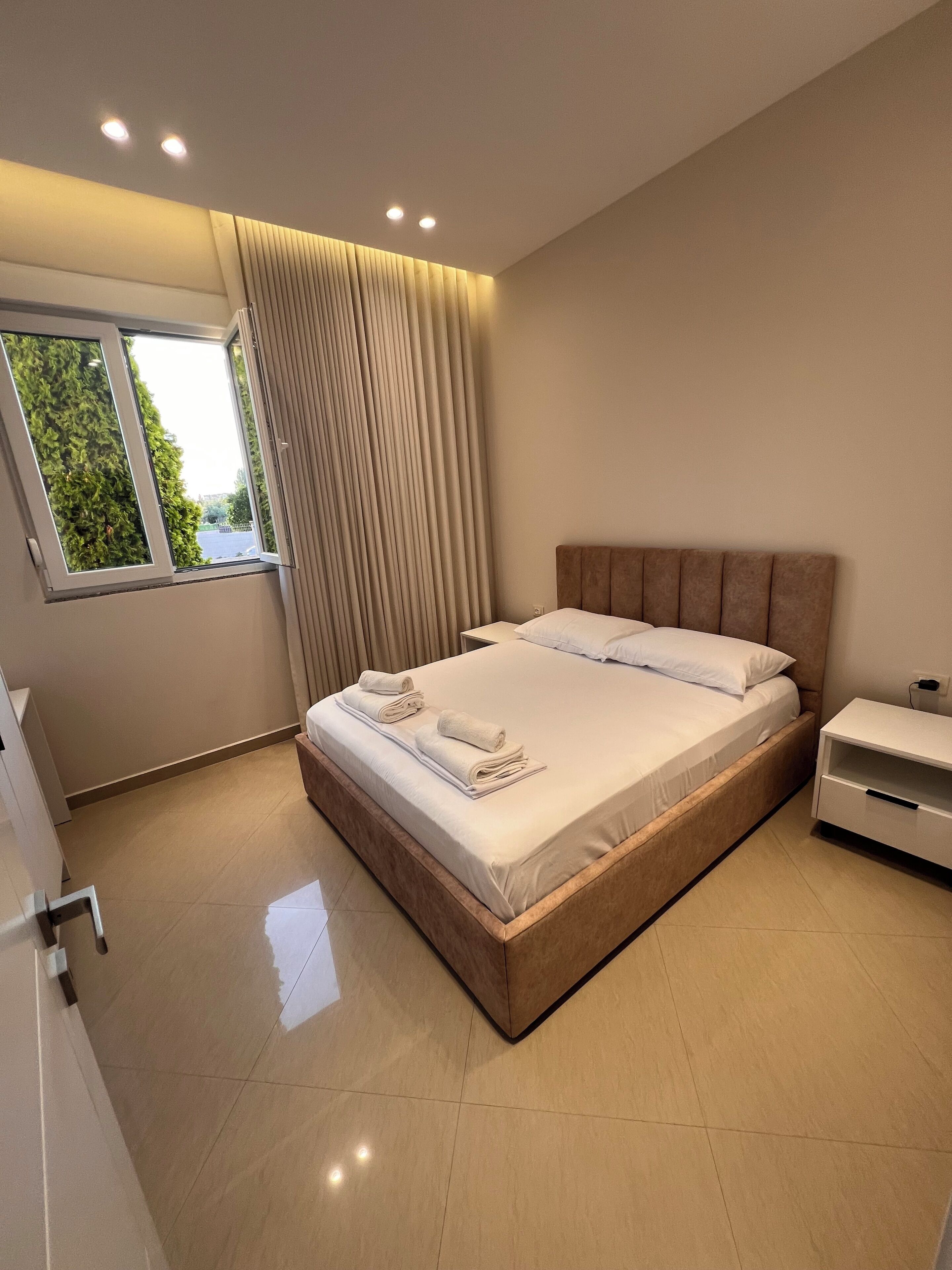 A modern bedroom with a large bed, a bedside table, a window with curtains, and a view of greenery outside.