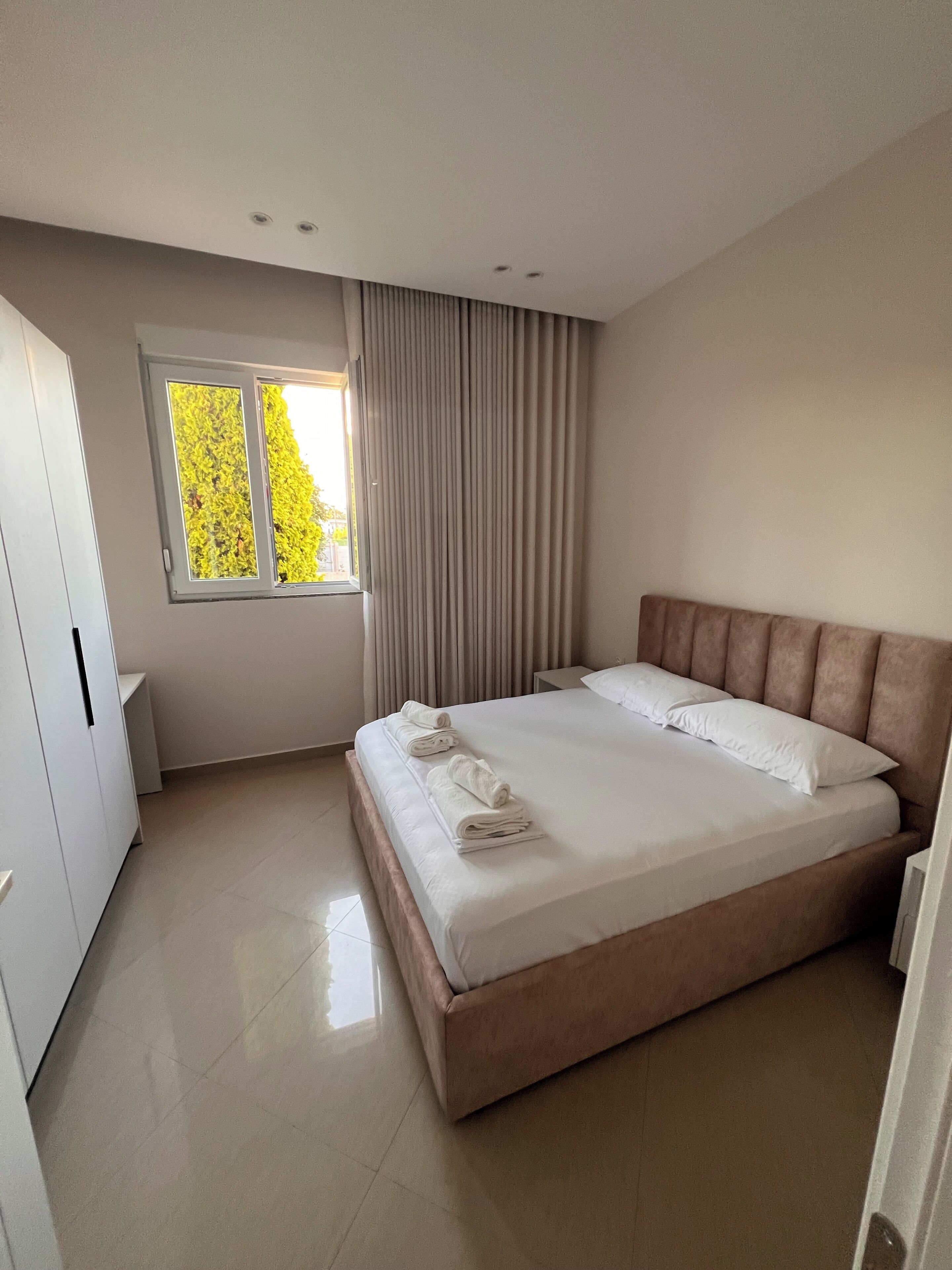 Een moderne hotelkamer met een groot bed, een raam met gordijnen, een kledingkast en een schone, betegelde vloer.