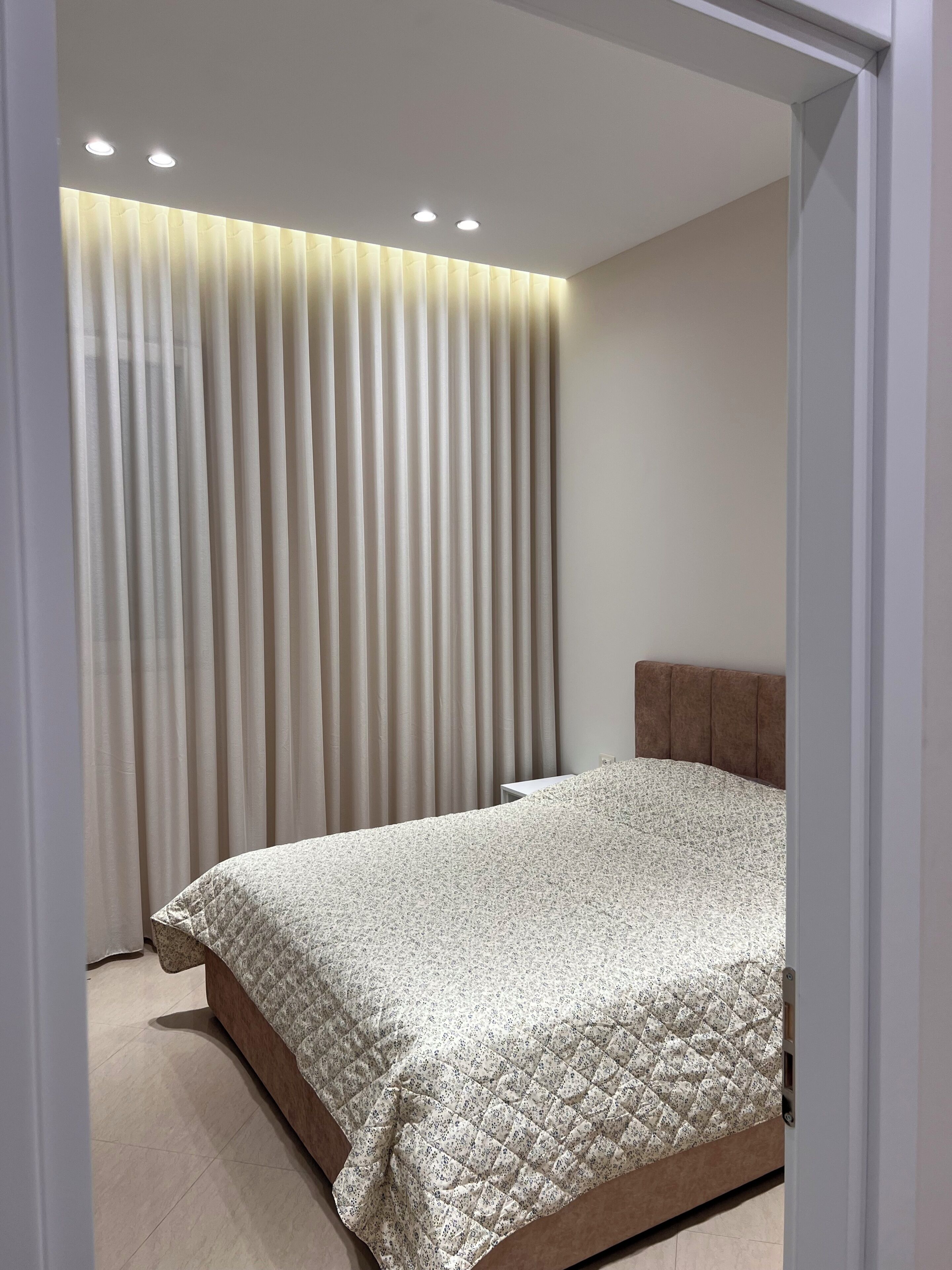 Una camera da letto con un letto, una testiera, una finestra con tende e faretti a soffitto.