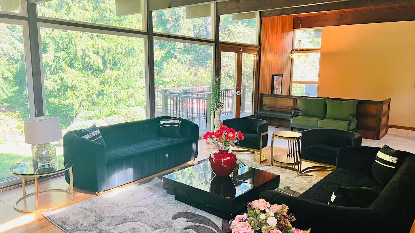 Una sala de estar moderna con ventanas grandes, un sofá verde, un sofá negro y una mesa de centro con flores.