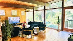 A modern living room with a television, black sofas, and a large window with a view of greenery. - Northwest Elegance – Modern Retreat (Normandy Park)