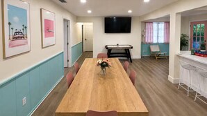 Un'area pranzo con un tavolo di legno, sedie rosa, un vaso di fiori, un tavolo da calcio e una TV montata a parete.