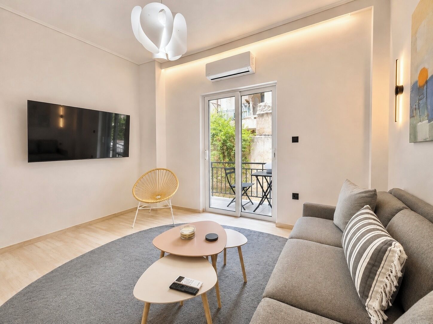 Ein modernes Wohnzimmer mit Flachbildfernseher, einem Rattan-Sessel, einem runden Couchtisch, einer Couch mit gestreiften Kissen und einer Schiebeglastür, die zu einem Außenbereich führt.