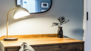 Ett träbord som står vid sängen, med en lampa, en vas med växter och en spegel som reflekterar ett fönster.