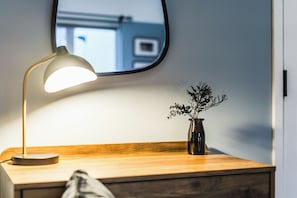 A wooden bedside table with a lamp, a vase with plants, and a mirror reflecting a window. - Crescent Apartments | Newport City Centre (Newport)