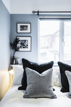 A bedroom with a bed, pillows, a nightstand, a lamp, framed pictures on the wall, and a window with a view. - Crescent Apartments | Newport City Centre (Newport)