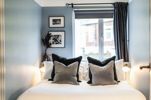 A neatly made bed with pillows, a window with curtains, and framed pictures on the wall. - Crescent Apartments | Newport City Centre (Newport)