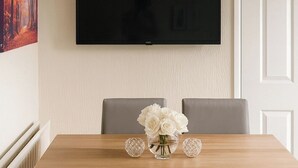 Ein Essbereich mit einem Holztisch, sechs grauen Stühlen, einer Vase mit weißen Blumen und einem an der Wand befestigten Flachbildfernseher.