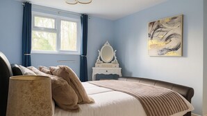 Ein Schlafzimmer mit einem Bett, einer Nachttischlampe, einem Schminktisch, einem Spiegel und einem Fenster mit Vorhängen.