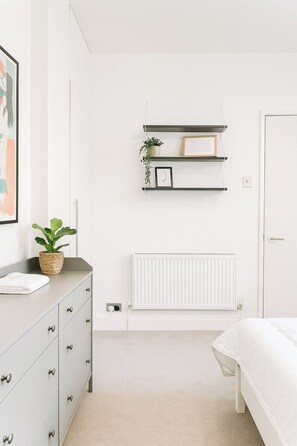 Ein Schlafzimmer mit Bett, Kommode, Heizkörper und Wandregalen mit Dekoration.