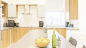 Une cuisine moderne avec des meubles en bois, un îlot central agrémenté d’un saladier de fruits et d’une bouteille, et une fenêtre avec des stores.