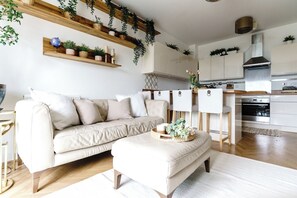 A modern living room with a sofa, ottoman, and a coffee table. There are wooden shelves with plants and a kitchen area with cabinets and a stove. - The Riverside Haven – City Centre + Parking (Newport)