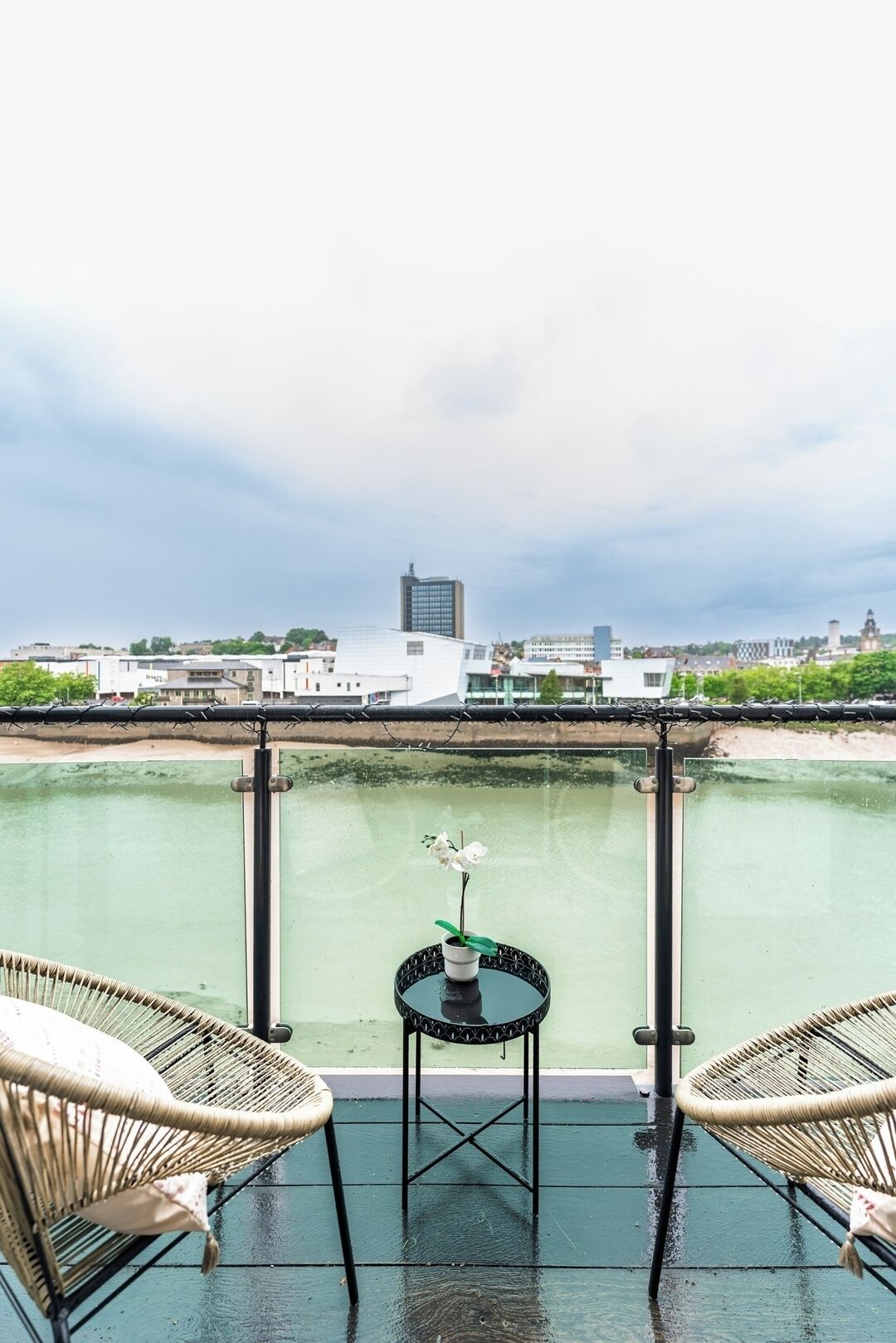 A balcony with two wicker chairs and a small table, overlooking a body of water and a cityscape.