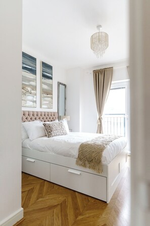 A bedroom with a tufted headboard, a bed with white bedding, a macrame throw, a chandelier, and a window with curtains. - The Riverside Haven – City Centre + Parking (Newport)