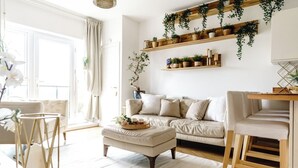 A living room with a sofa, ottoman, wooden shelves with plants, and a dining area with a chair. - The Riverside Haven – City Centre + Parking (Newport)