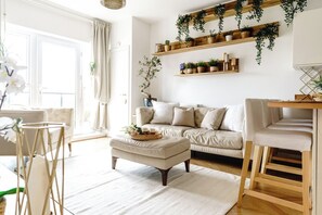 Ein Wohnzimmer mit einer Couch, einem Hocker, Holzregalen mit Pflanzen und einem Essbereich mit einem Stuhl.