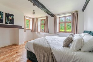 A bedroom with a large bed, wooden floor, two windows with curtains, and a hanging light fixture. - 1641 Historic Farmhouse, Sleeps 16 · Bath/Bristol (Keynsham)