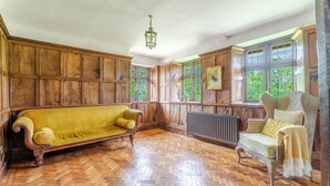 Ein Zimmer mit Holzvertäfelung, einem Fischgrätparkett, einem gelben Sofa, einem weißen Sessel und einem Heizkörper.