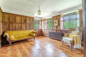 A room with wooden paneled walls, a herringbone wooden floor, a yellow sofa, a white armchair, and a radiator. - 1641 Historic Farmhouse, Sleeps 16 · Bath/Bristol (Keynsham)
