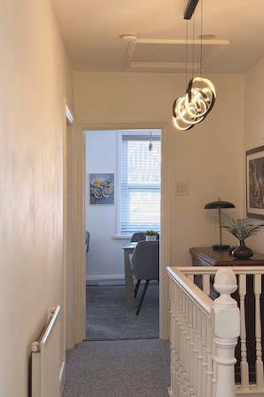 A hallway with a staircase leading to a room with a desk, chair, and a window with blinds. - The M4 Escape | J26 | Free Parking | Newport (Newport)