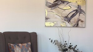 Ein brauner Sessel mit gemustertem Kissen, ein großes abstraktes Bild an der Wand und eine Vase mit weißen Blumen auf einem Teppichboden.
