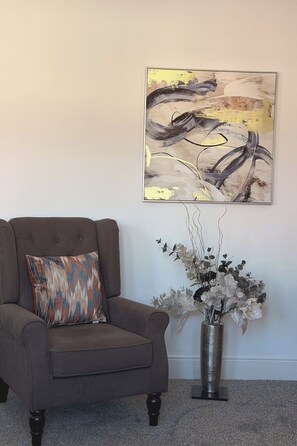 A brown armchair with a patterned cushion, a large abstract painting on the wall, and a vase with white flowers on a carpeted floor. - The M4 Escape | J26 | Free Parking | Newport (Newport)