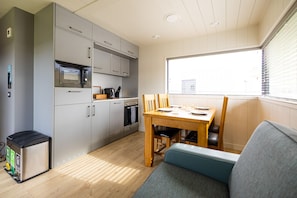 A compact kitchen with a microwave, oven, and sink, a dining table with chairs, and a window with blinds. - Lost Shore Surf Resort (Newbridge)